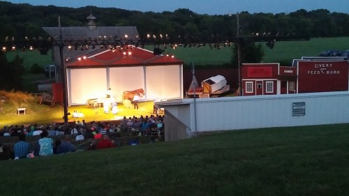 A theater performance outdoors with a horse, set against a backdrop of buildings and a large audience in the foreground.
