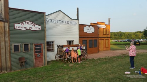 A western-style town with wooden buildings, a wagon, and people posing for a photo in a grassy area.