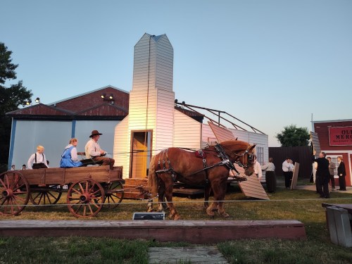 A horse-drawn wagon in front of a partially constructed building with actors in period costumes during an outdoor performance.