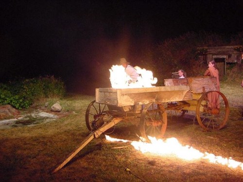 A wooden cart on fire at night, with people nearby watching the flames and smoke in a rural setting.