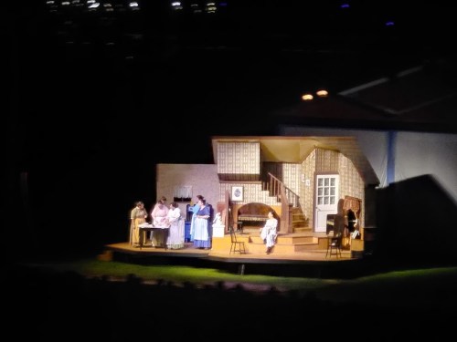 A stage scene depicting a domestic setting with actors in period costumes engaged in various activities.
