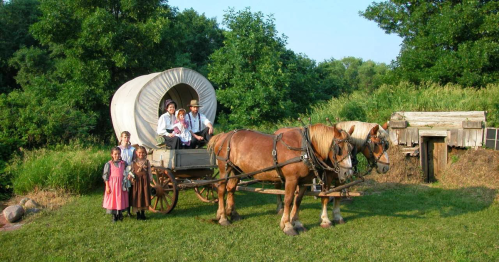 A horse-drawn wagon with people in traditional attire, surrounded by greenery and a rustic setting.