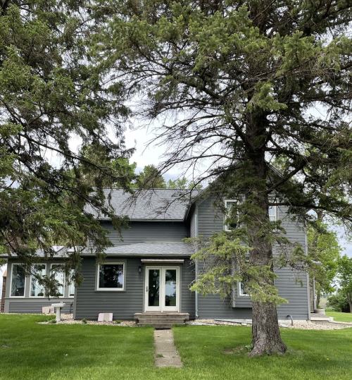 A gray two-story house surrounded by green grass and trees, featuring a front porch and large windows.