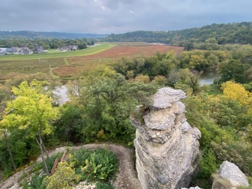 A rocky outcrop overlooks a lush valley with a winding river and colorful autumn foliage under a cloudy sky.