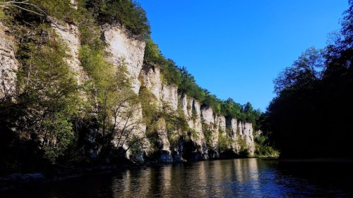 A serene river scene with towering cliffs and lush greenery under a clear blue sky.