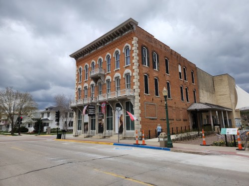 Historic brick building with decorative balconies, flags, and cloudy sky in a small town setting.