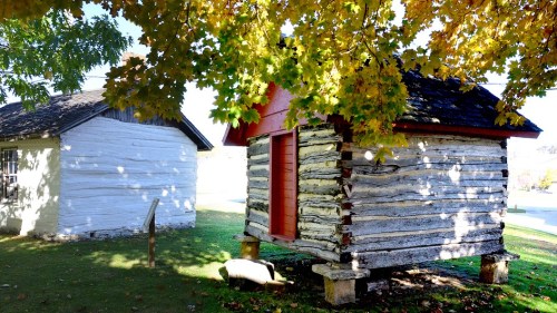 Two rustic log cabins surrounded by vibrant autumn leaves, with a clear blue sky in the background.