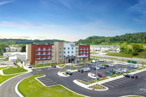 A modern hotel with a parking lot, surrounded by greenery and other buildings under a clear blue sky.