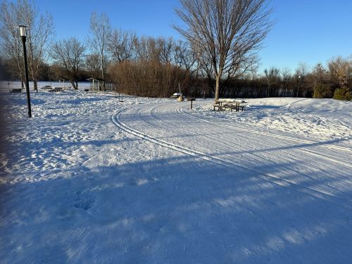 A snowy landscape with a winding path, trees, and a picnic area under a clear blue sky.