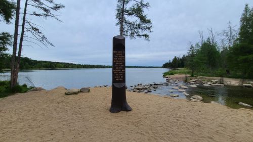 A wooden monument stands on a sandy shore by a calm river, surrounded by trees and rocky banks under a cloudy sky.