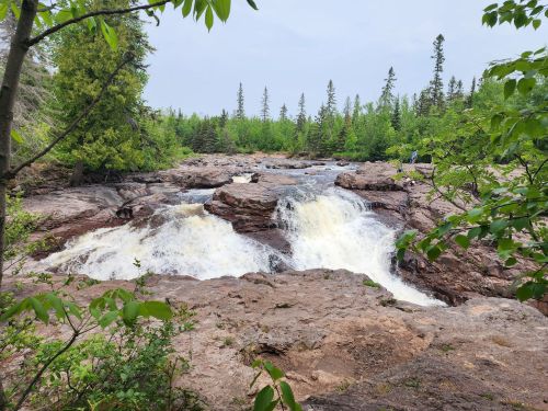 A scenic view of a waterfall cascading over rocky terrain, surrounded by lush green trees and a cloudy sky.