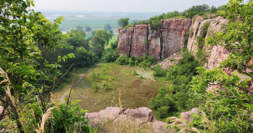 A rocky cliff surrounded by greenery, overlooking a grassy area and distant fields under a clear sky.