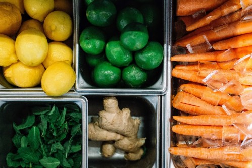 A variety of fresh produce: lemons, limes, ginger, mint, and carrots arranged in metal containers.