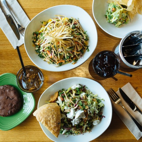A top-down view of a table with salads, a cookie, chips, and drinks on a wooden surface.