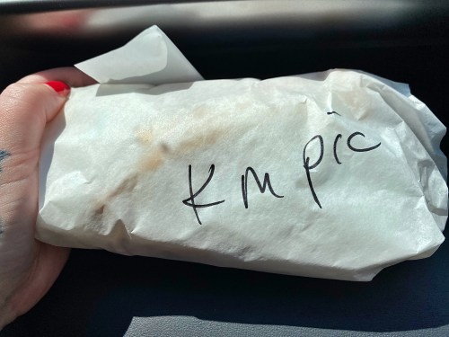 A hand holding a wrapped food item with handwritten text "KM pic" on the paper.