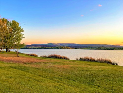 A serene lakeside view at sunset, featuring green grass, trees, and distant mountains under a colorful sky.