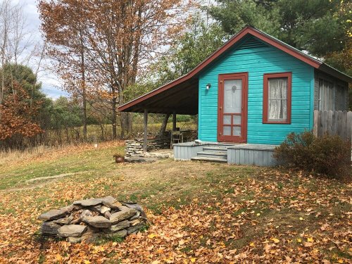 A small turquoise cabin with a porch, surrounded by autumn leaves and a stone fire pit in the foreground.