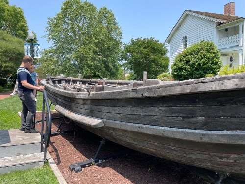 Two people stand beside a weathered wooden boat on display, with a historic house and greenery in the background.