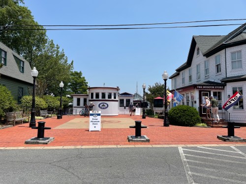 A sunny plaza with a circular building, benches, and shops, featuring a sign that says "Enter Here."