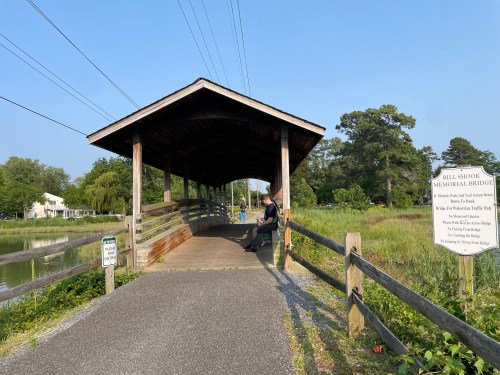 A covered wooden bridge over a calm waterway, with a sign about the bridge and a person sitting nearby.