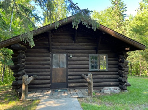 A rustic log cabin surrounded by trees, featuring a wooden porch and a single door with a window.