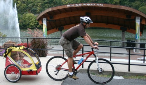 A man rides a bike with a child trailer past Hazel Ruby McQuain Park, with a fountain and lake in the background.