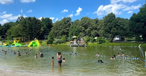 A lively outdoor swimming area with people enjoying the water, surrounded by trees and a sunny sky.