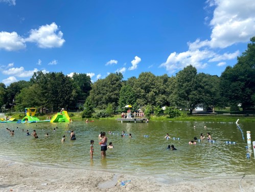 A sunny day at a lake with people swimming, a water slide, and trees in the background.
