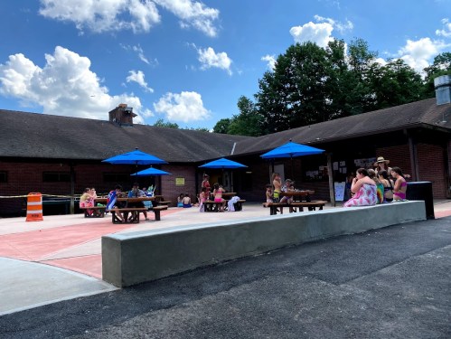 A sunny outdoor area with people sitting at tables under blue umbrellas, near a brick building and trees.