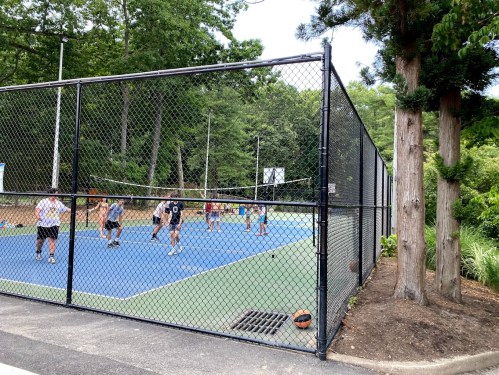 A fenced tennis court with players engaged in a match, surrounded by trees and greenery.