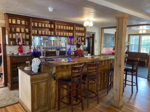 A cozy bar with wooden decor, beer taps, and a bartender, featuring shelves of mugs and colorful lights.