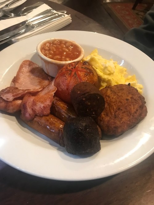 A plate of breakfast featuring scrambled eggs, sausages, bacon, a grilled tomato, baked beans, and a black pudding slice.