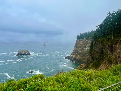 A misty coastal scene featuring cliffs, rocky outcrops, and waves crashing against the shore under a cloudy sky.