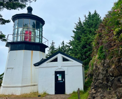 A white lighthouse with a glass lantern room, surrounded by trees and rocky terrain under a cloudy sky.