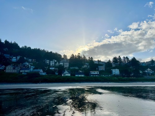 Sunrise over a coastal town, with houses on a hillside and reflections on the wet sand of the beach.
