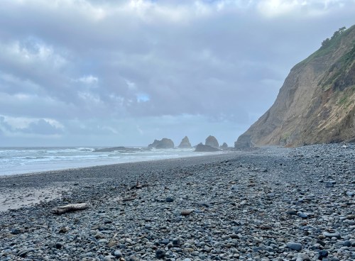 A rocky beach with smooth stones, waves crashing, and cliffs under a cloudy sky.