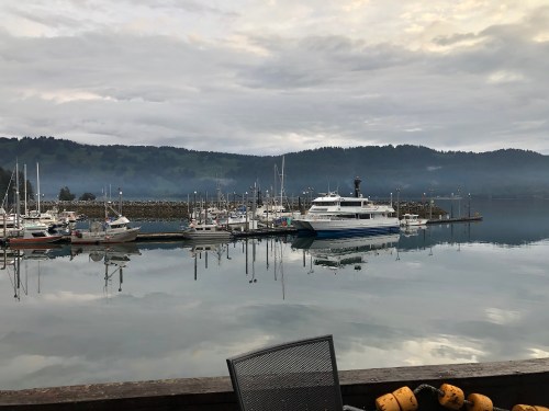 A calm marina with boats docked, reflecting in the water under a cloudy sky and misty mountains in the background.