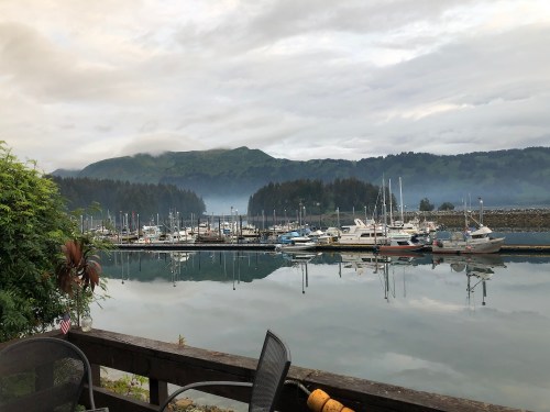 A serene marina with boats docked, reflecting mountains and cloudy skies in calm water.