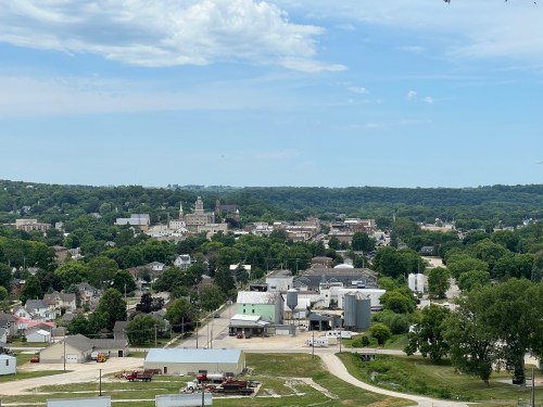 A panoramic view of a small town surrounded by greenery, featuring buildings and a clear blue sky.