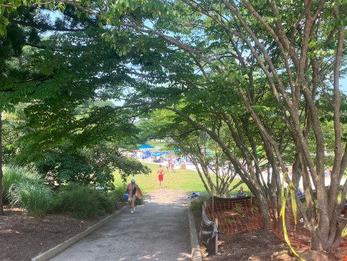 A pathway lined with trees leads to a pool area, with people enjoying the sun in the background.