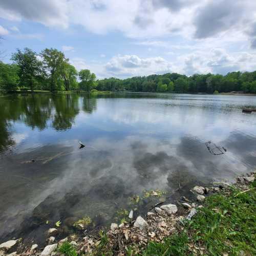 A serene lake surrounded by greenery, reflecting clouds and trees under a partly cloudy sky.