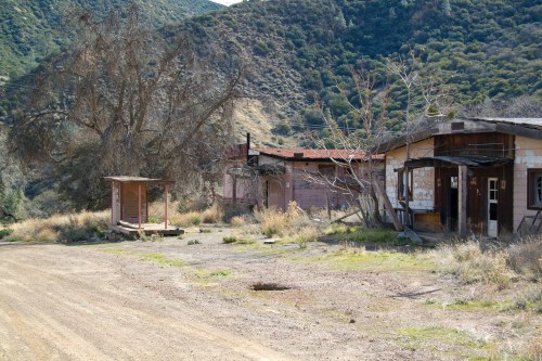 Abandoned buildings along a dirt road, surrounded by dry grass and hills in the background.