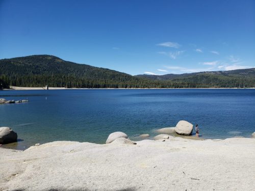 A serene lake surrounded by mountains, with a person standing on a rocky shore under a clear blue sky.
