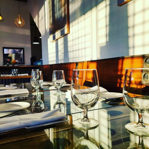 A dining table set with glassware and plates, illuminated by soft shadows from nearby windows.