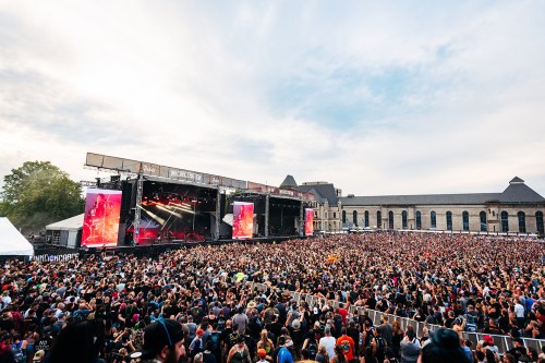 A large crowd at an outdoor music festival, with a stage and colorful lights under a cloudy sky.