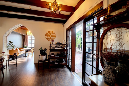 Cozy interior of a cafe with wooden beams, plants, and a decorative wall, featuring large windows and warm lighting.