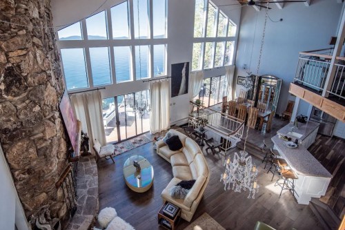 Spacious living room with large windows, stone fireplace, and modern decor overlooking a serene lake view.