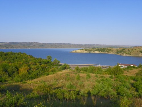 A serene lake surrounded by green hills under a clear blue sky, with a small house near the water's edge.