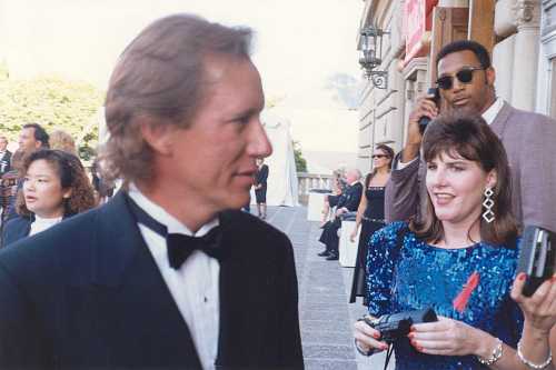 A man in a tuxedo walks past a woman in a sequined dress, while another man in the background talks on a phone.