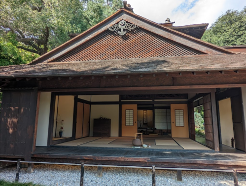 Traditional Japanese house with wooden architecture, sliding doors, and a tatami mat interior, surrounded by greenery.
